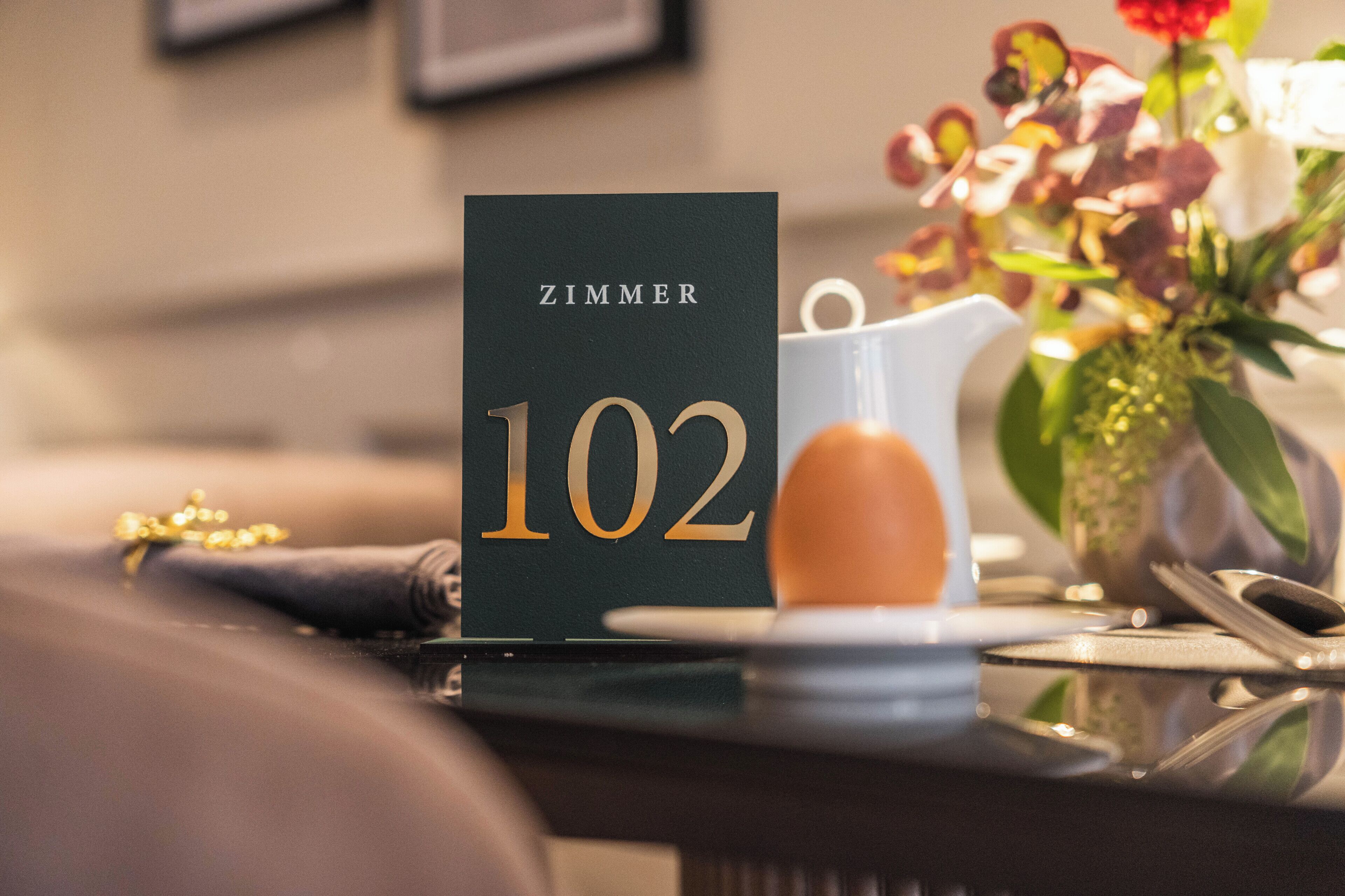 A hotel room number sign, a white pitcher, and a vase with flowers on a table.