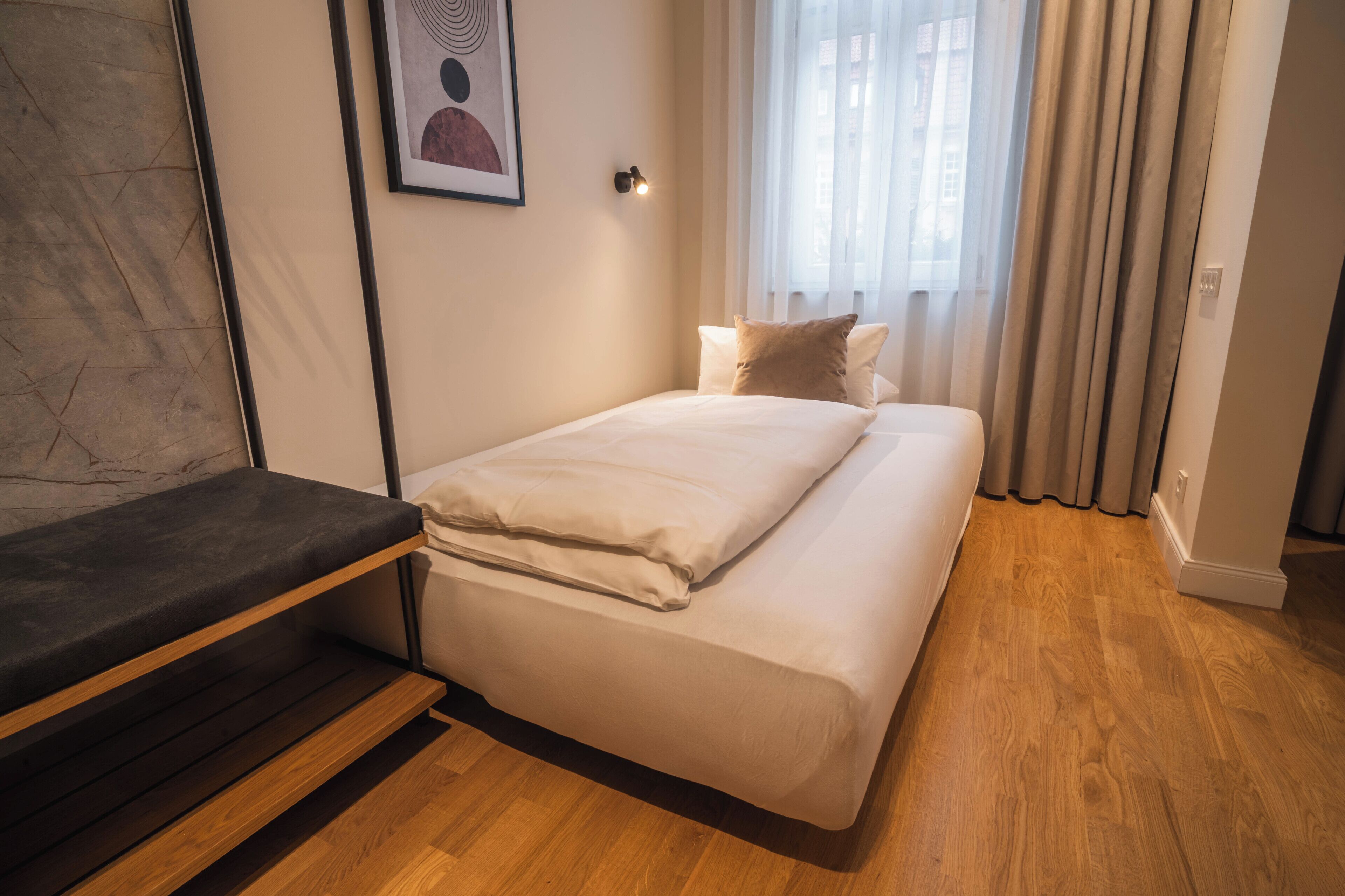 A bedroom with a bed, a bench, a wall-mounted light, and a window with curtains.