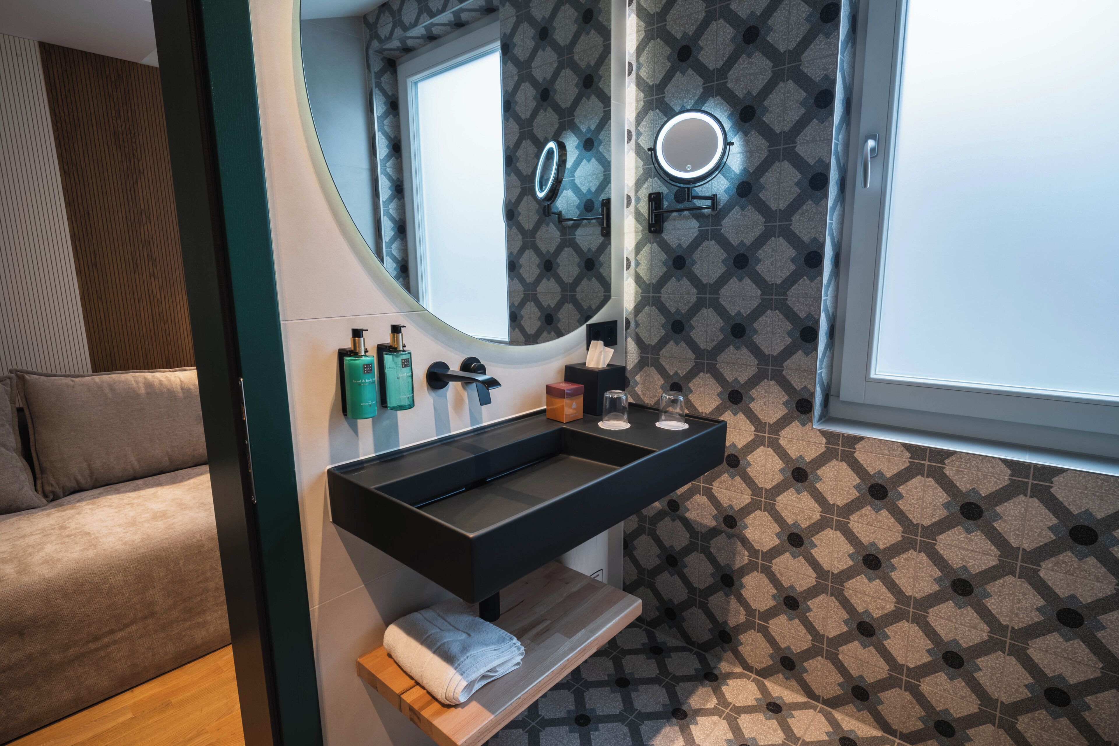 Un baño moderno con un lavabo negro, dos espejos, un perchero para toallas y baldosas estampadas.