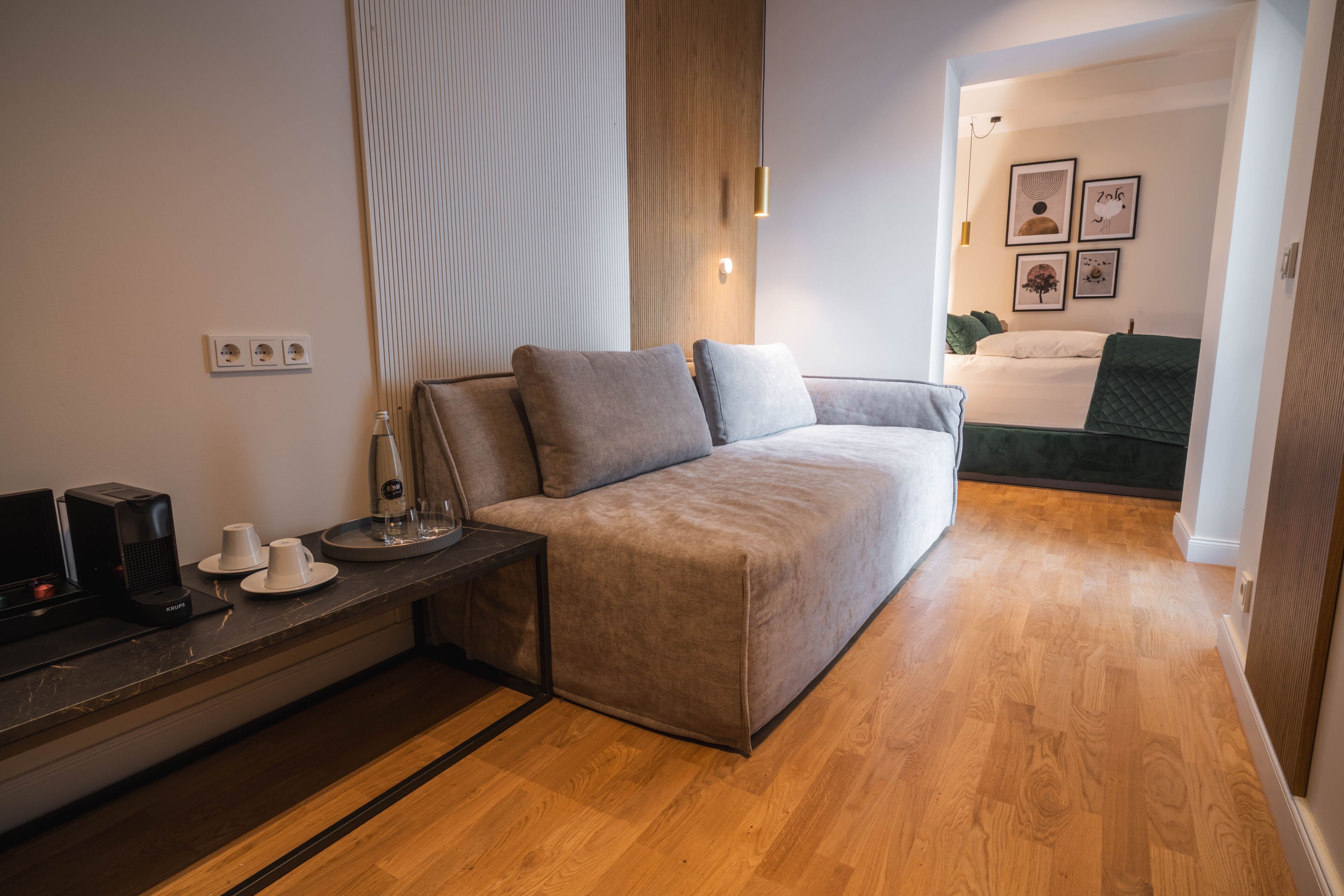 Una habitación moderna con sofá, una mesita de centro, televisión y escritorio. Se ve una cama con una manta verde al fondo.