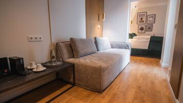 Una habitación moderna con sofá, una mesita de centro, televisión y escritorio. Se ve una cama con una manta verde al fondo.