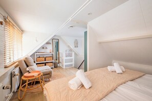 Ein Schlafzimmer mit einem Bett, einem Sessel, einem kleinen Tisch, einem Regal und einem Fenster mit Jalousien.