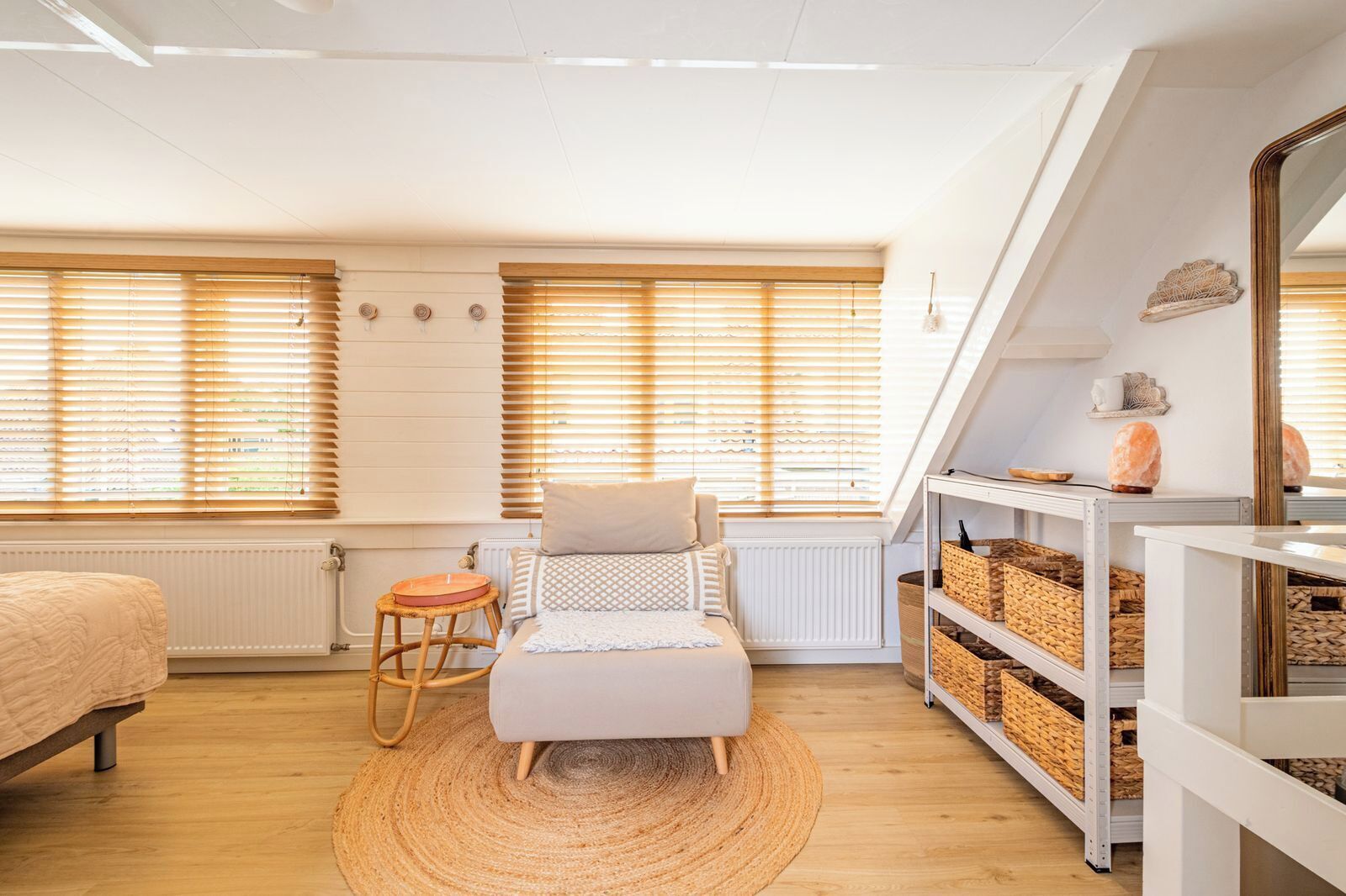 A cozy room with a bed, a small seating area, a shelf with baskets, and a large window with blinds.