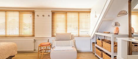 Una habitación acogedora con una cama, un pequeño rincón de descanso, una estantería con cestas y un amplio ventanal con persianas.