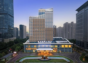 Un hotel moderno con un destacado edificio CMOC al fondo, que cuenta con un amplio muro de cristal y una terraza en la azotea.