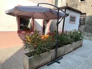 Una jardinera de concreto con diversas plantas y flores, una sombrilla de patio y un pequeño edificio con ventana.