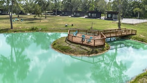A lakeside deck with chairs and a wooden bridge connecting to a small island. - Relaxing Tiny Home Retreat with Pool, Goats & Firepits near Houston in Hempstead, Texas (Hempstead)