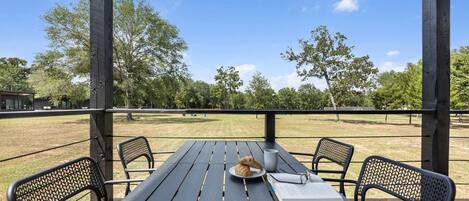 An outdoor wooden table with chairs, a dog, and a book on a deck with a view of a grassy field and trees.