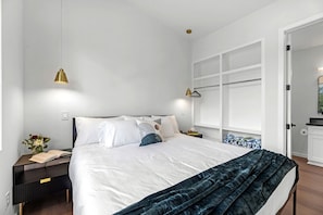 Une chambre moderne avec un grand lit, des tables de chevet, une commode, une fenêtre avec des stores et une porte donnant sur une autre pièce.