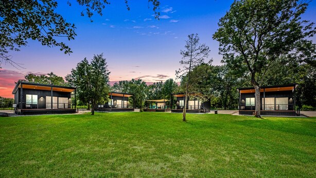 A modern building complex with multiple structures, surrounded by a well-maintained lawn and trees. - Tranquil Tiny Home Resort with Heated Pool & Goats near Houston, Hempstead Texas (Hempstead)