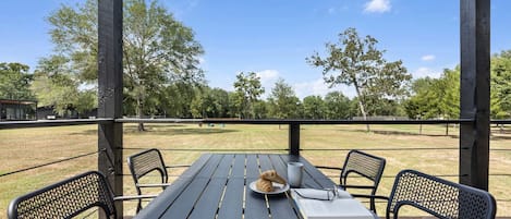 Una mesa de madera al aire libre con sillas, un perro y un libro en una terraza con vistas a un campo de césped y árboles.