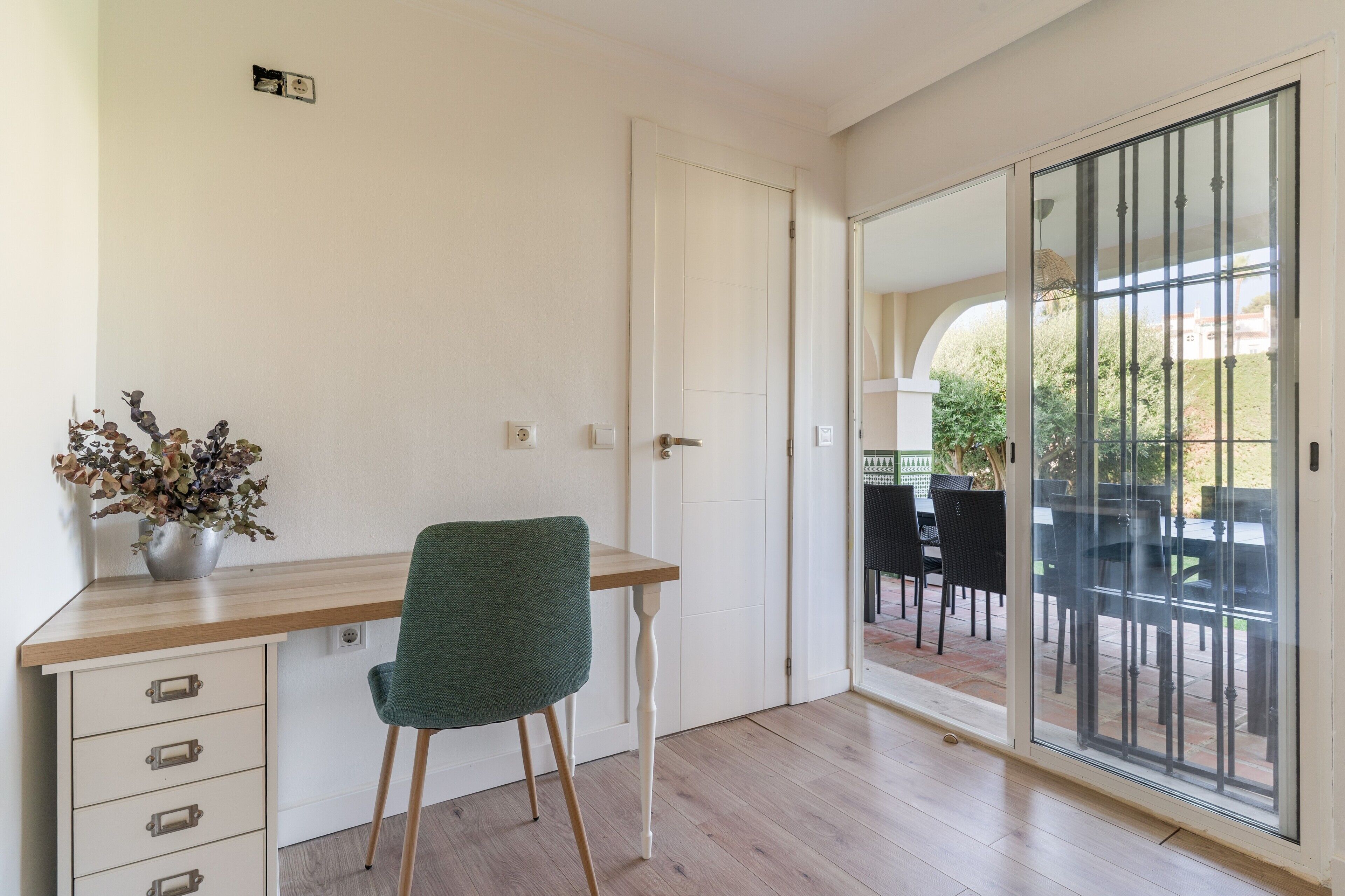 Une pièce avec un bureau en bois, une chaise verte, une plante en pot et une porte-fenêtre coulissante donnant sur un patio extérieur.
