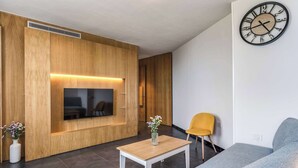 A modern living room with a flat-screen TV, wooden panel walls, a grey sofa, a yellow chair, and a wall clock. - Sea View Suite Priora with Private Terrace (Priora)