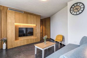A modern living room with a flat-screen TV, wooden panel walls, a grey sofa, a yellow chair, and a wall clock. - Sea View Suite Priora with Private Terrace (Priora)