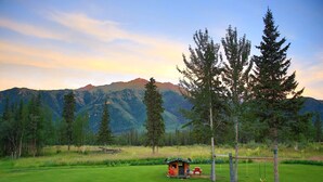 A grassy field with a small wooden structure, trees, and mountains in the background. - Historic Cozy Off-Grid Cabin with Creekside Steam Sauna near Log Cabin Creek, Tok, Alaska (Tok)