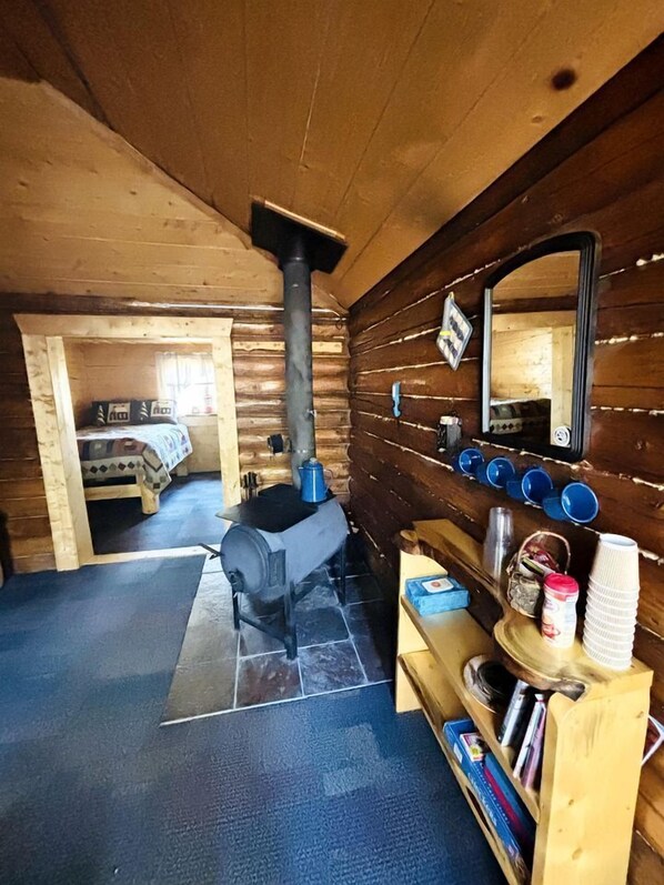 A log cabin interior with a wood-burning stove, a bed, a mirror, and a shelf with books and cups.
