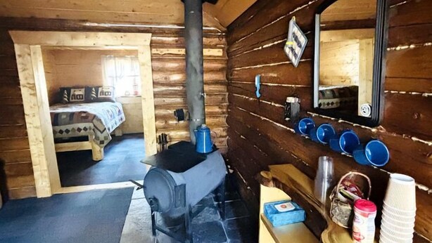 A log cabin interior with a wood-burning stove, a bed, a mirror, and a shelf with books and cups. - Historic Cozy Off-Grid Cabin with Creekside Steam Sauna near Log Cabin Creek, Tok, Alaska (Tok)