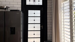 A hallway with a black door featuring a geometric pattern, a trash bin, and a window with blinds. - Herbert House (Cape Town)