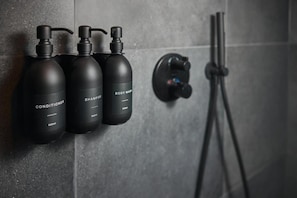 Un baño con tres botellas negras etiquetadas 'CONDITIONER', 'SHAMPOO' y 'BODY WASH', y un cabezal de ducha.