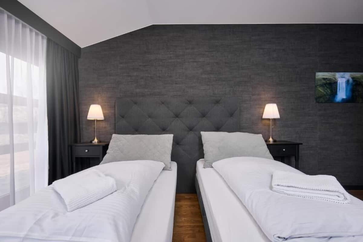 A hotel room with two beds, a grey upholstered headboard, a painting on the wall, and a window with curtains.
