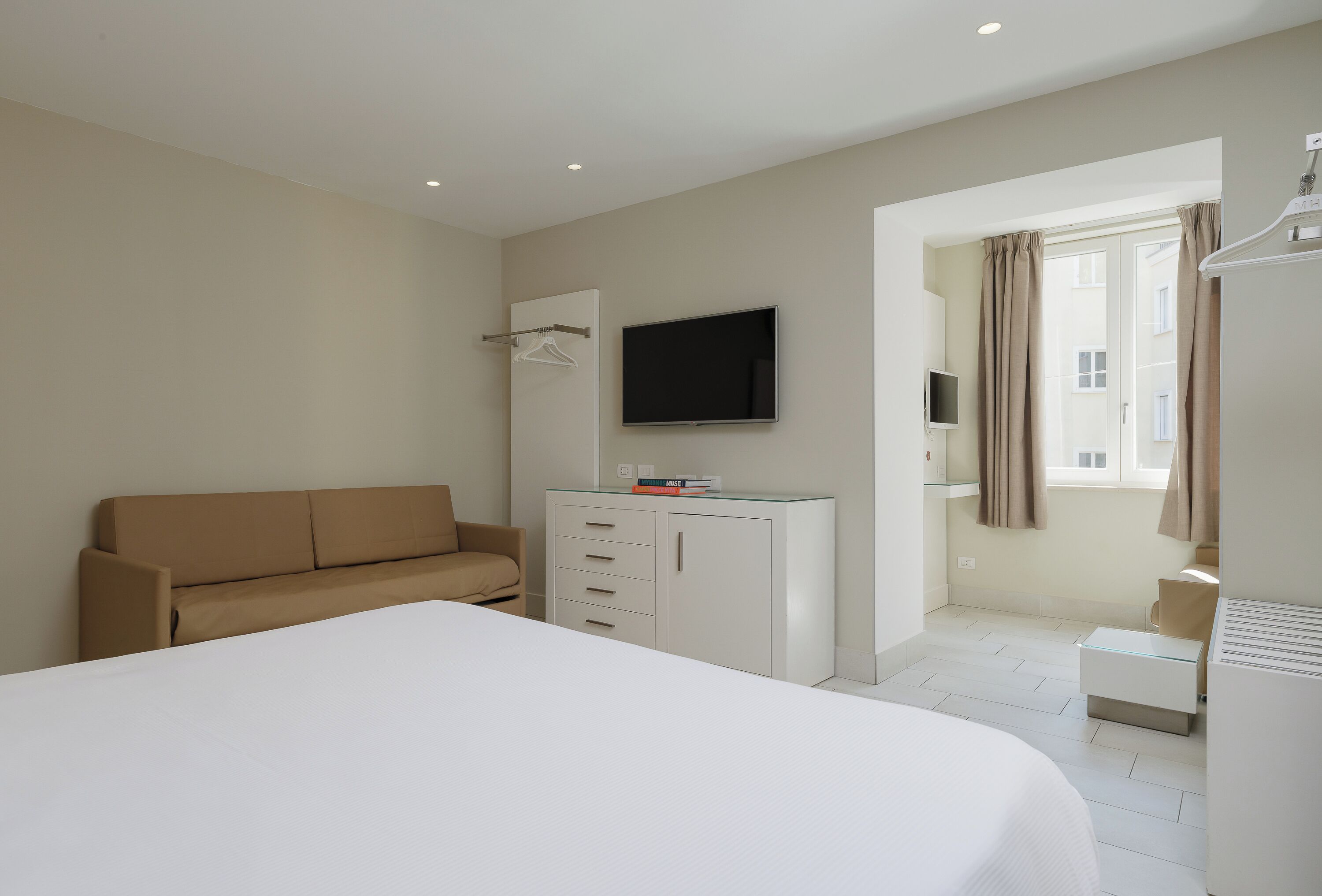 A hotel room with a bed, a sofa, a TV mounted on the wall, a small desk, and a window with curtains.