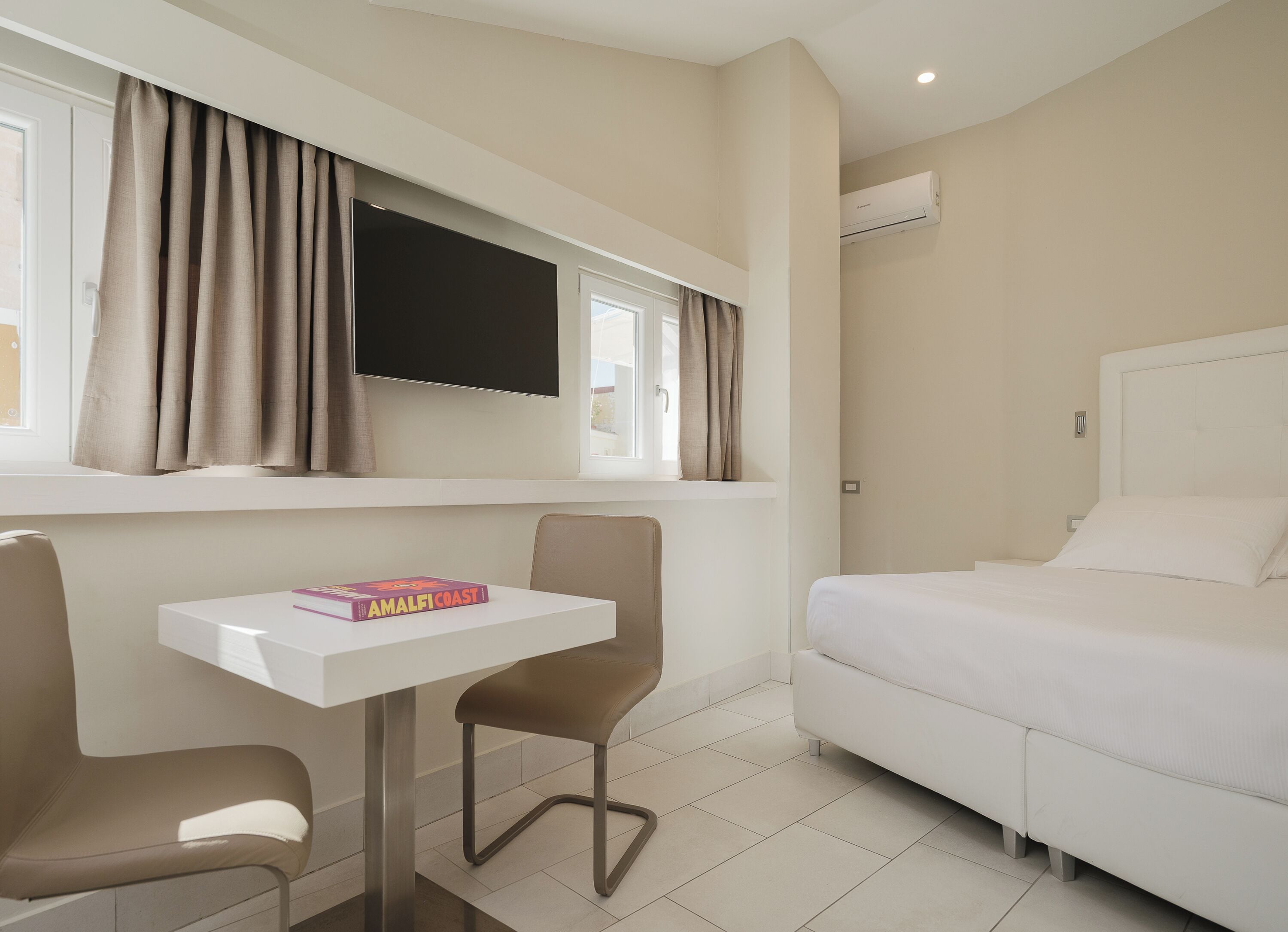 Une chambre d’hôtel avec un lit, un téléviseur fixé au mur, une petite table avec une chaise et une fenêtre avec des rideaux.