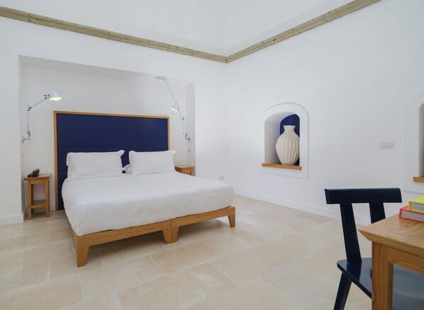 A bedroom with a bed, bedside tables, a nightstand, a chair, a small table, and a wall-mounted shelf with a vase.