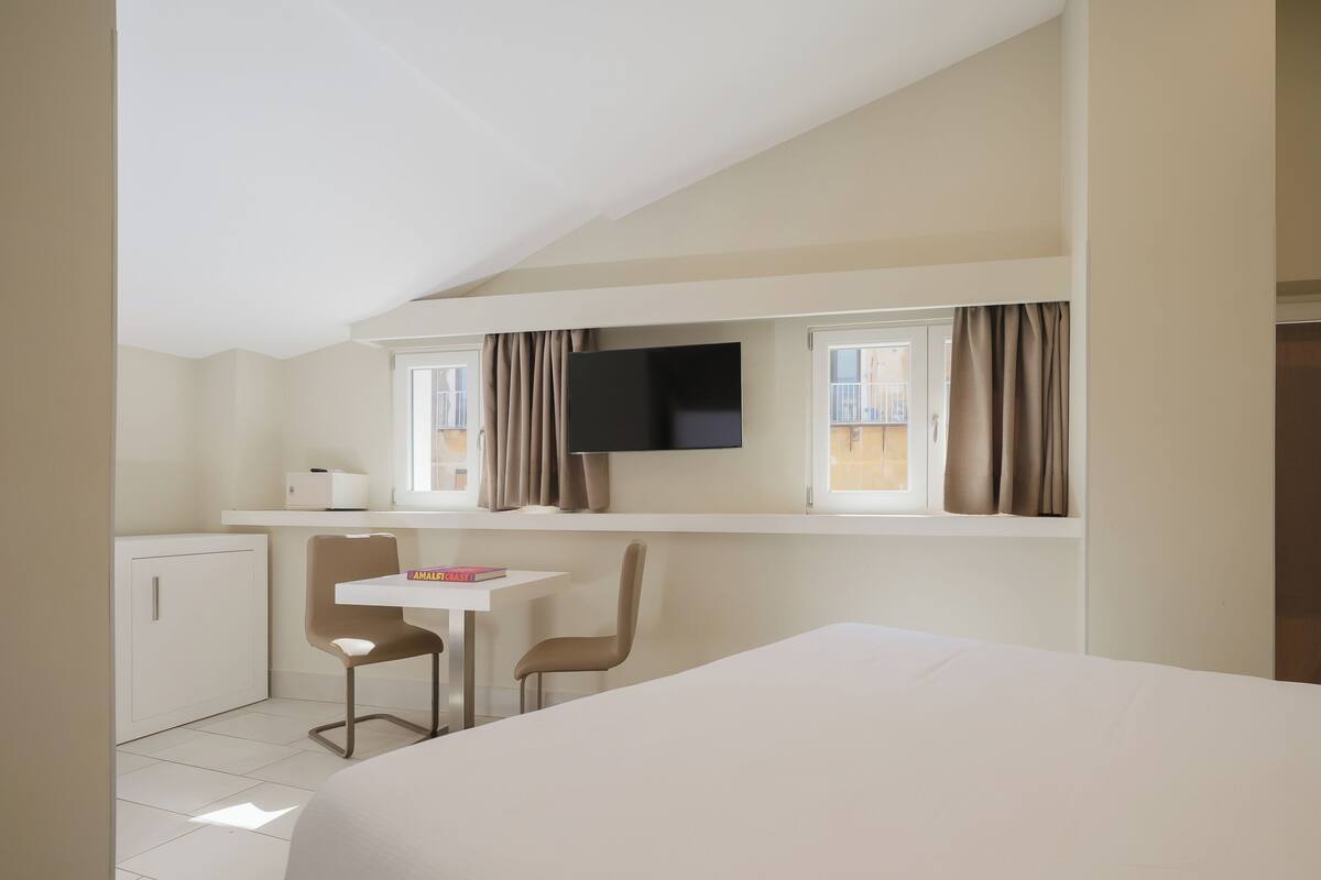 A bedroom with a bed, a desk with two chairs, a TV, and a window with curtains.