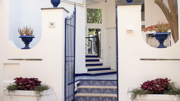 Un edificio blanco con detalles azules, que incluye una escalera con azulejos estampados, un cartel con el número '56' y plantas en macetas.