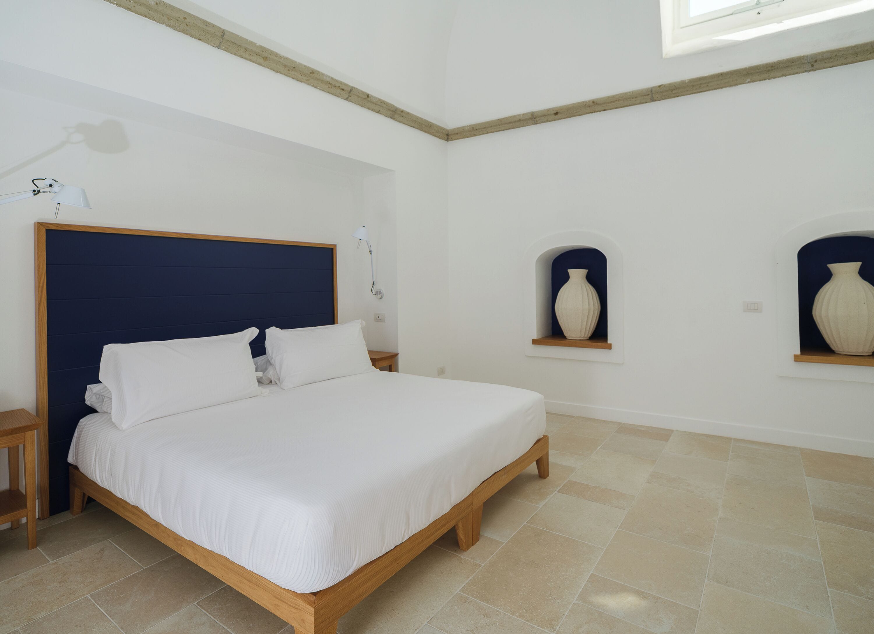Une chambre à coucher avec un lit en bois, du linge de lit blanc, une table de chevet, une lampe fixée au mur et deux étagères murales ornées de vases.