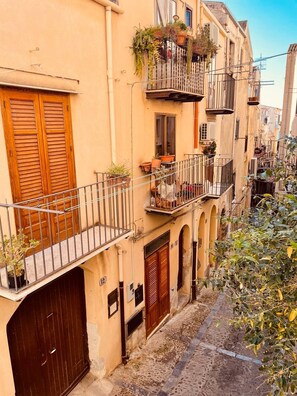 A narrow street with buildings featuring balconies with potted plants, wooden shutters, and a cobblestone path. - Romantic Cefalù - Studio Apartment in the Heart of the Historic Center (Cefalù)