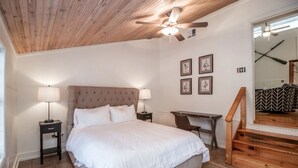 Ein Schlafzimmer mit einem Bett, zwei Nachttischlampen, einem Schreibtisch, einem Stuhl, einem Deckenventilator und einer Treppe.