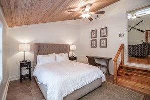 Ein Schlafzimmer mit einem Bett, zwei Nachttischlampen, einem Schreibtisch, einem Stuhl, einem Deckenventilator und einer Treppe.
