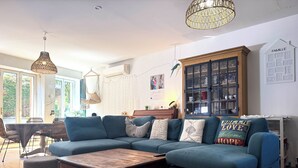 A living room with a sectional sofa, a coffee table, a hanging light fixture, a cabinet with glass doors, and a wall-mounted air conditioning unit. - Air-conditioned family home with a pool near Lyon (Sathonay-Camp)