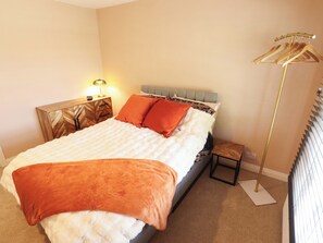 Ein ordentlich bezogenes Bett mit weißer und orangefarbener Decke, ein Nachttisch aus Holz mit einer Lampe und eine Stehlampe.