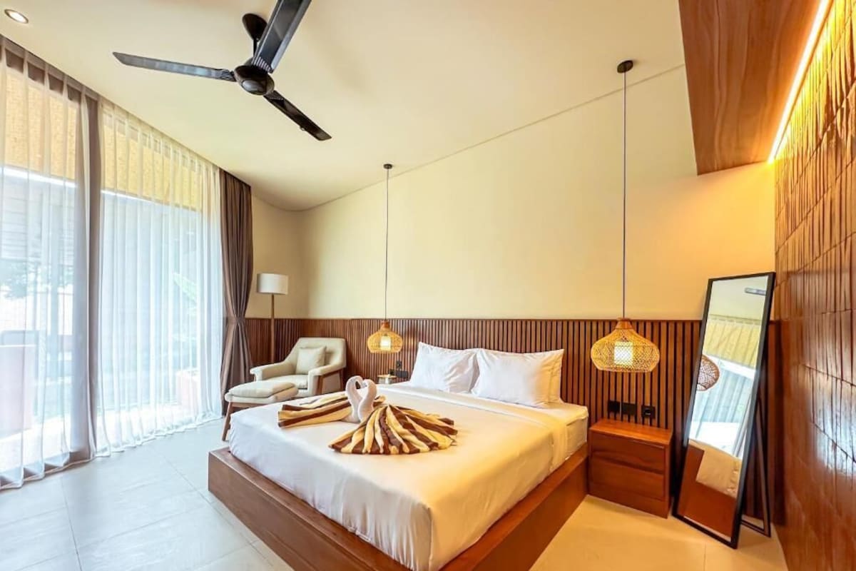A hotel room with a large bed, a ceiling fan, a nightstand, a lamp, and a view of the outdoors.