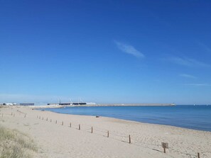 A beach with a fence, a clear sky, and a distant port. - Exclusive Apartment - Playa de Marenys de Rafaelcaid (Gandia)