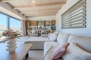 A modern living room with a sofa, a dining area with chairs, and a kitchen with wooden cabinets and a large window. - Rodia Luxury villa ,Sea View Retreat near Plakias (Plakias)