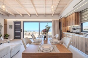 A modern kitchen with wooden cabinets, a dining area with a table set for four, and a view of the beach through the sliding glass door. - Rodia Luxury villa ,Sea View Retreat near Plakias (Plakias)