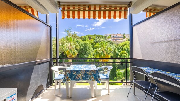 Un balcón con una mesa y sillas, vistas a zonas verdes y edificios, y un toldo a rayas.