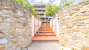 Un pasillo de paredes de piedra que conduce a una escalera con una puerta metálica, flanqueado por vegetación y un edificio moderno con balcones.