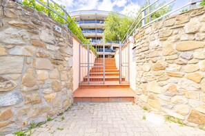 A stone-walled corridor leading to a staircase with a metal gate, flanked by greenery and a modern building with balconies. - Le Palme by Wonderful Italy (Sanremo)