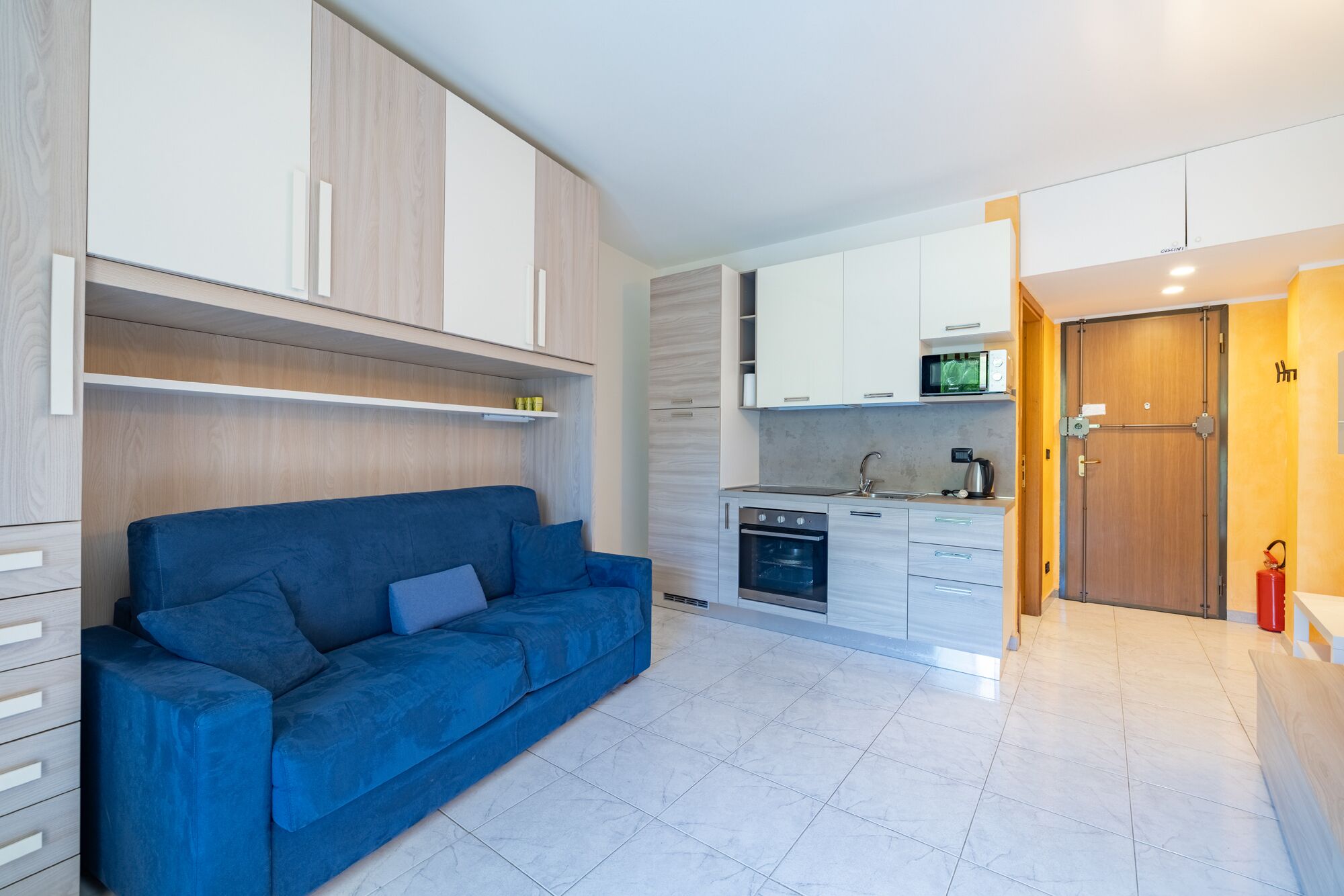 Een kleine woonruimte met een bank, kitchenette en een deur naar een andere kamer.