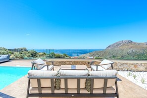 Ein Poolbereich mit einer Couch und Sesseln, einer Steinmauer und Blick auf das Meer und die Hügel.
