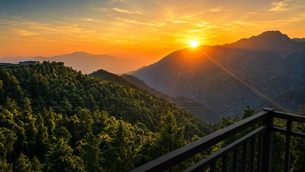 Paisaje montañoso con un atardecer, una baranda de madera y un edificio distante.