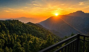 Paisagem montanhosa com pôr do sol, um corrimão de madeira e um edifício ao longe.