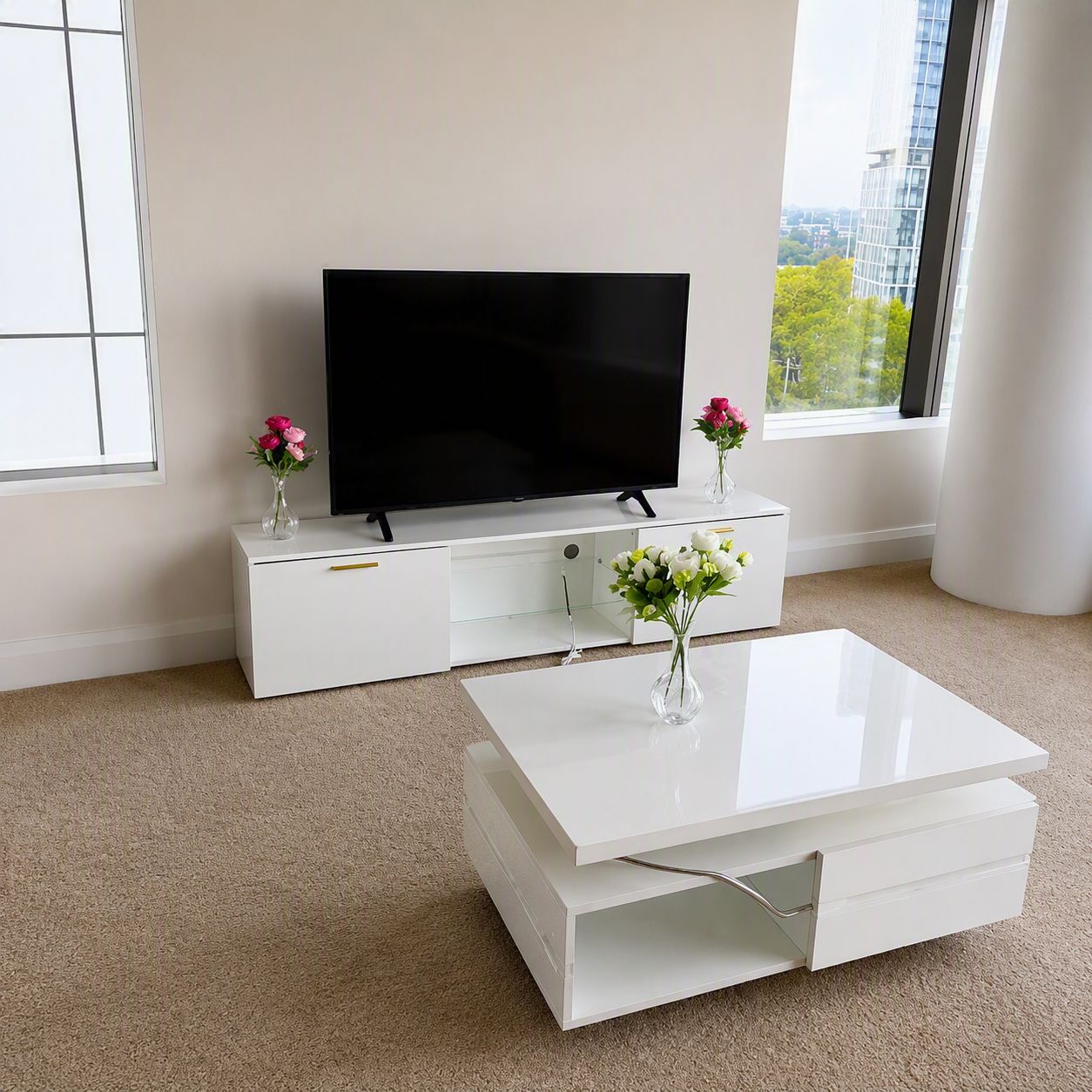 A modern living room with a flat-screen TV on a white entertainment unit, a white coffee table with a vase of flowers, and a large window offering a view of buildings and trees.