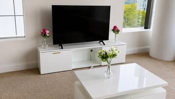 A modern living room with a flat-screen TV on a white entertainment unit, a white coffee table with a vase of flowers, and a large window offering a view of buildings and trees.