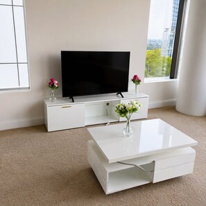 A modern living room with a flat-screen TV on a white entertainment unit, a white coffee table with a vase of flowers, and a large window offering a view of buildings and trees. - Stylish Apartment Sydney Olympic Park Free parking (Sydney Olympic Park)