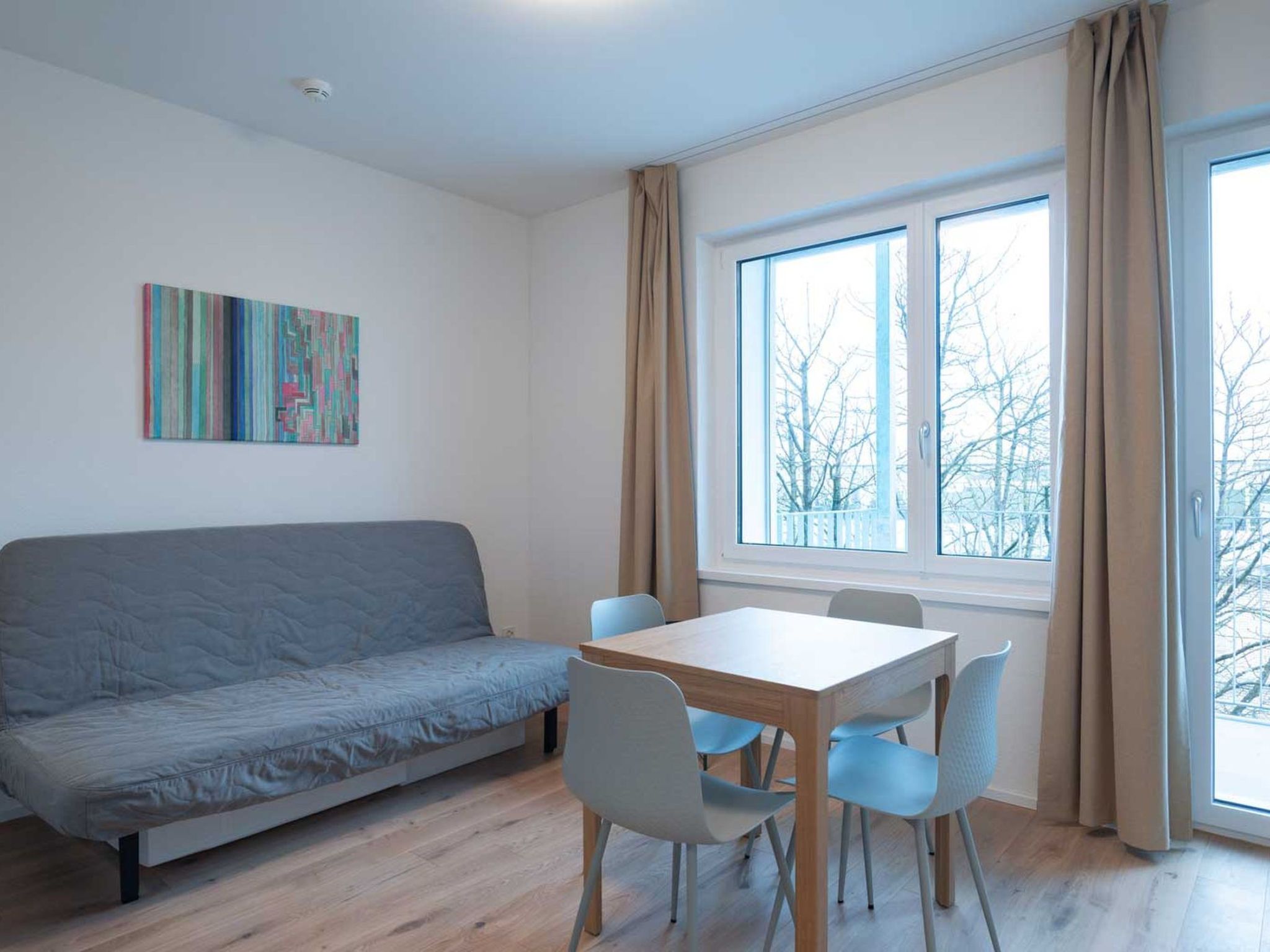 A room with a grey sofa, a wooden table with chairs, a window with curtains, and a ceiling light.